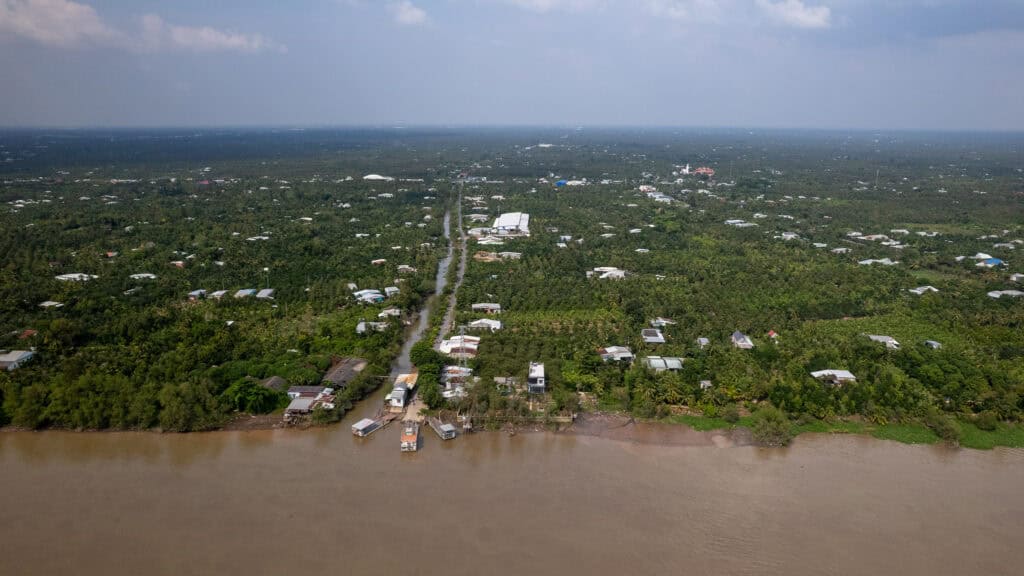 Aerial view of a rural riverside village, perfect for a Vietnam to Cambodia tour, with scattered houses, green trees, and a road extending from the river into the forested landscape under a partly cloudy sky.