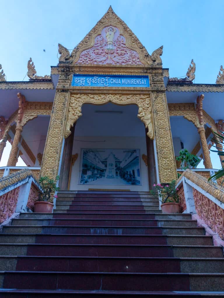 Grand entrance of Chua Munirensay, adorned with ornate gold carvings, two staircases with potted plants, and blue sky in the background—an inspiring sight often admired on many Vietnam to Cambodia tours. If you’re wondering how to get from Vietnam to Cambodia, this magnificent temple stands as a memorable stop along the popular Ho Chi Minh City to Phnom Penh route.