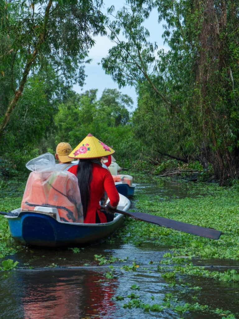 Two people paddle a boat through a lush, green, tree-filled waterway on a scenic vietnam to cambodia tour. The person in front wears a conical hat and red clothing, surrounded by floating vegetation and calm water under a blue sky.