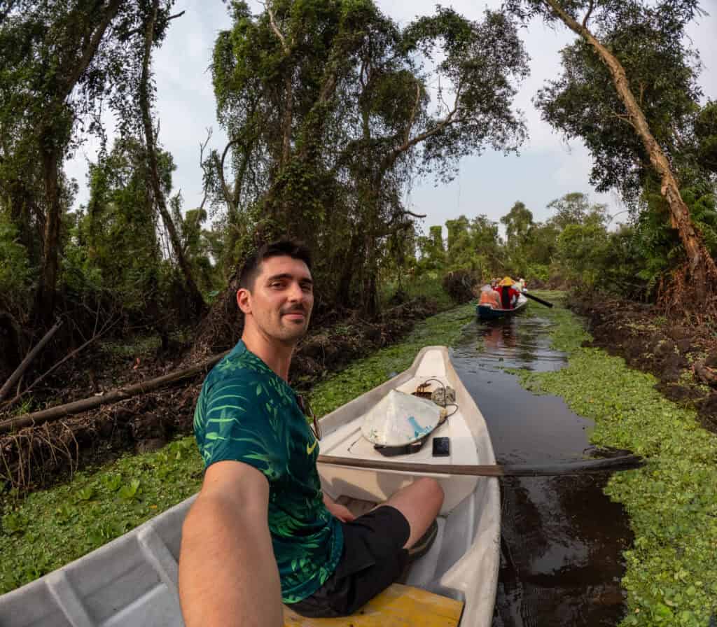 A man takes a selfie while paddling a canoe through a narrow, plant-filled waterway surrounded by lush green trees—just one of the scenic moments you might experience on a Vietnam to Cambodia tour; another canoe is visible ahead, highlighting the sense of adventure on the journey from Ho Chi Minh City to Phnom Penh.