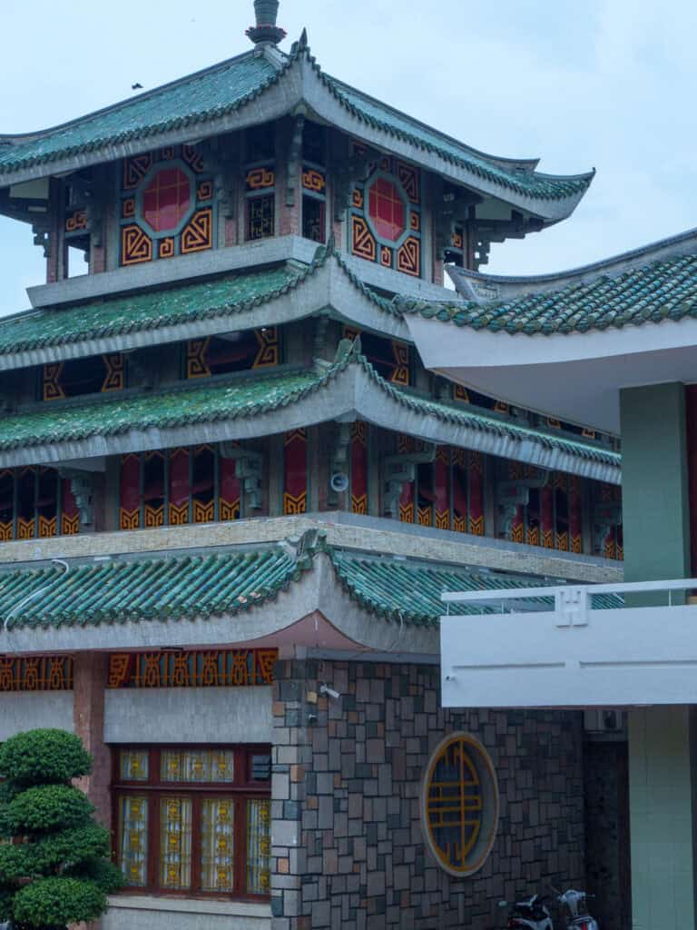 A multi-story building with traditional Asian architectural elements, green tiled roofs, red decorative windows, and stone walls stands adjacent to a modern white structure—reminiscent of the vibrant contrasts you might see when traveling from Ho Chi Minh City to Phnom Penh along the popular route from Vietnam to Cambodia.
