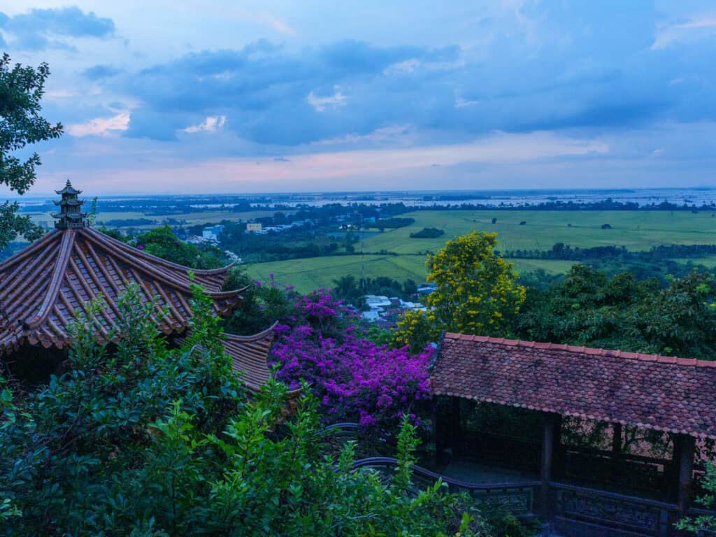 A scenic view of tiled-roof pavilions surrounded by lush greenery and purple flowers, overlooking vast green fields and a distant river under a blue, cloudy sky at sunset—perfectly capturing the enchanting landscapes you can experience on a Vietnam Cambodia tour from Ho Chi Minh to Phnom Penh.