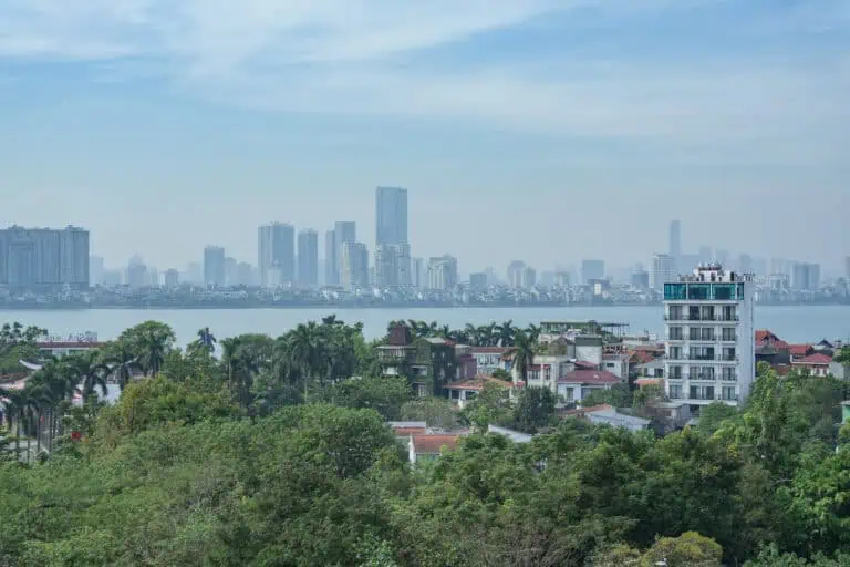 A cityscape with tall buildings in the background, a body of water in the middle, and houses surrounded by trees and greenery in the foreground under a partly cloudy sky—this picturesque scene evokes some of the top things to visit in Hanoi.