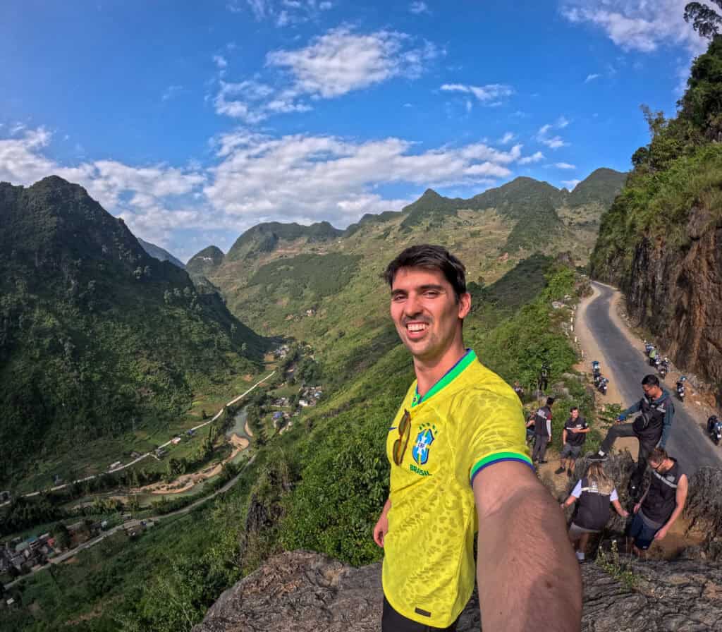 A man in a yellow jersey takes a selfie on a mountain road along the famous Ha Giang Loop, with green valleys, winding road, blue sky, and a group of people with motorbikes in the background.