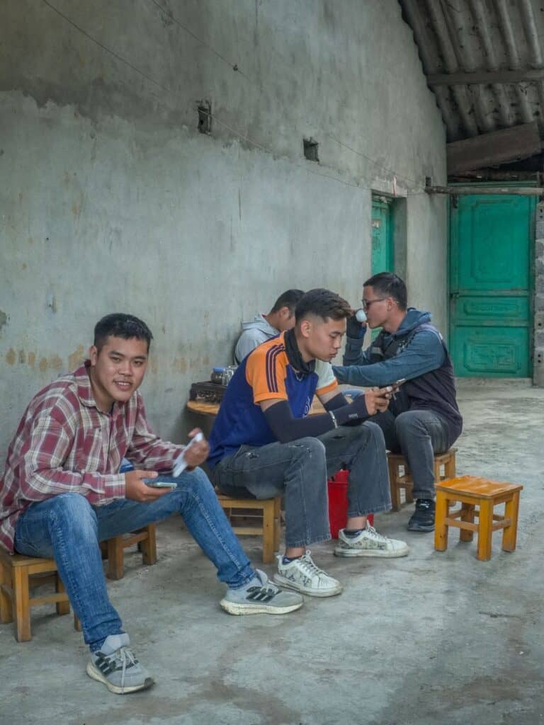 Four young men sit on small wooden stools by a worn concrete wall, with green doors in the background—a scene that captures everyday life along the Ha Giang Loop Vietnam. Two are focused on their phones, while one in a plaid shirt smiles at the camera, perhaps taking a break from planning their Ha Giang Loop itinerary.