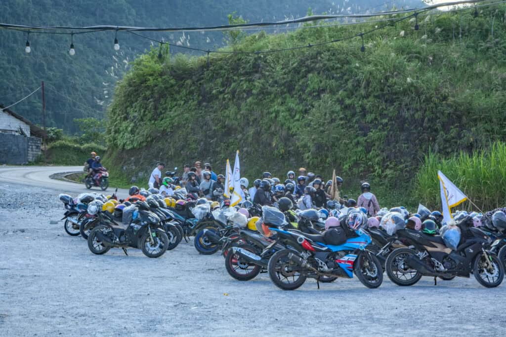 A group of motorcyclists gathers beside a row of parked motorcycles on a gravel road along the stunning Ha Giang Loop, surrounded by greenery and hills, with string lights overhead.