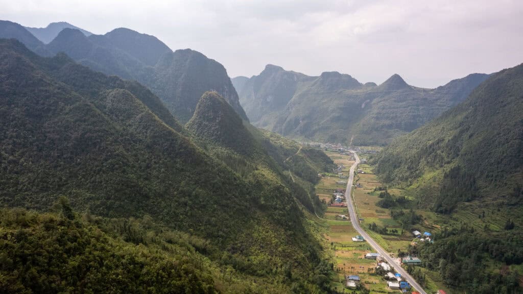 A winding road runs through a lush green valley surrounded by tall mountains, with scattered houses and fields along the route under a cloudy sky, capturing the breathtaking scenery of the Ha Giang Loop.