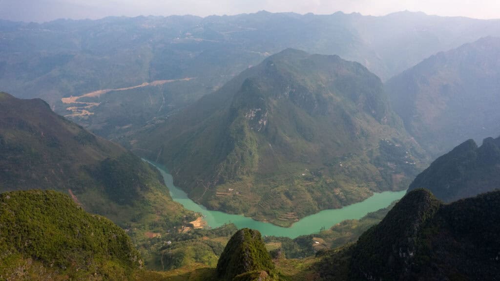 A winding turquoise river flows through a lush green valley surrounded by steep mountains under a hazy sky. Small settlements dot the landscape at the rivers edge.
