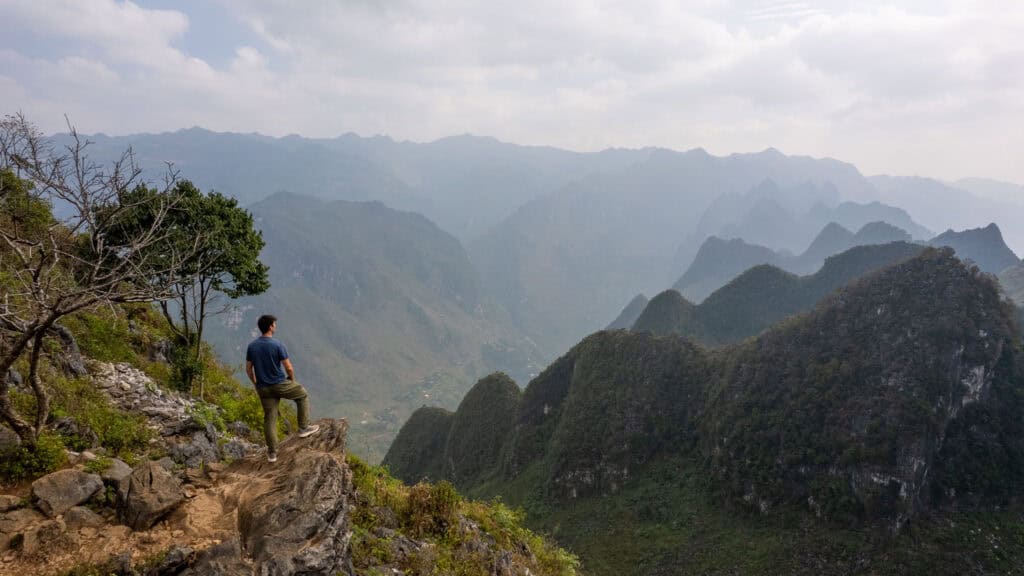 A person stands on a rocky ledge, looking out over a scenic valley filled with lush, green mountains under a cloudy sky.