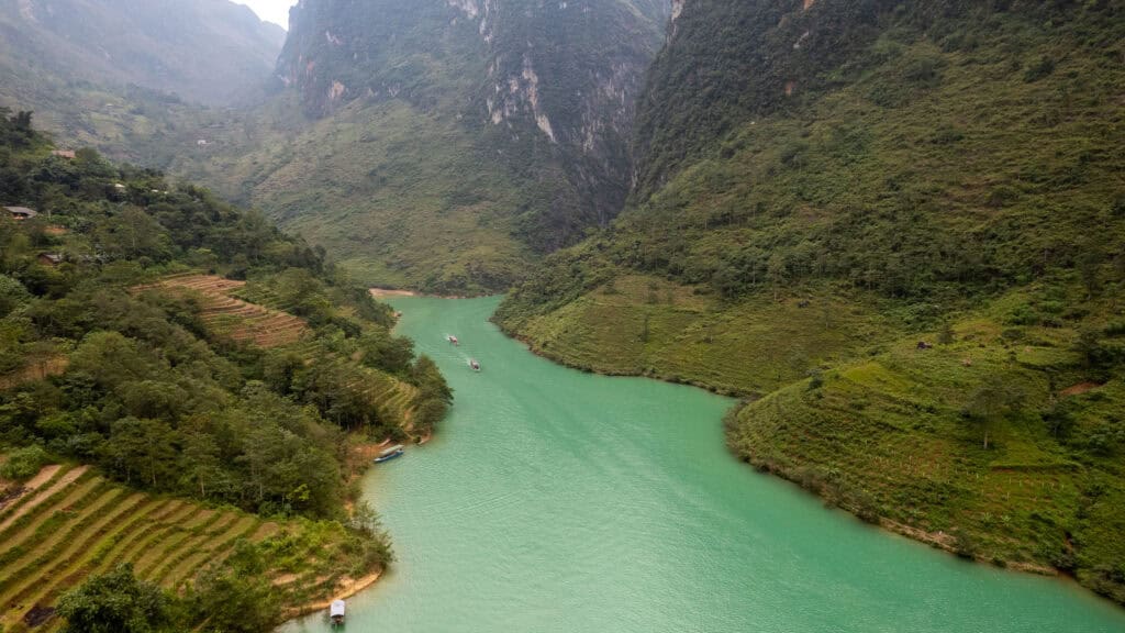 A turquoise river winds through lush green mountains and terraced hillsides, with boats visible on the water under a misty sky.
