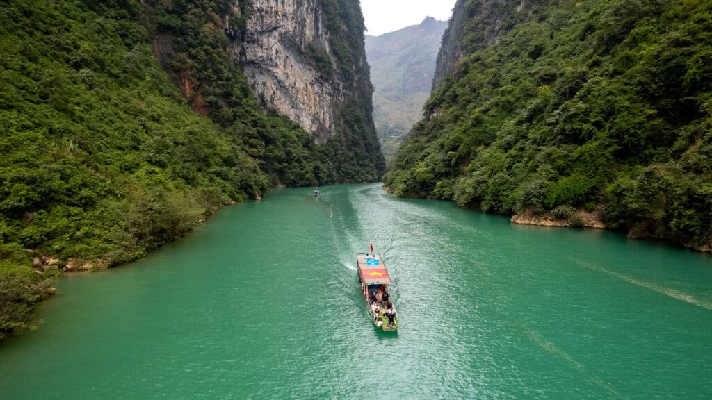 A boat with people travels along a turquoise river flanked by steep, green cliffs under a cloudy sky, with distant mountains visible in the background.