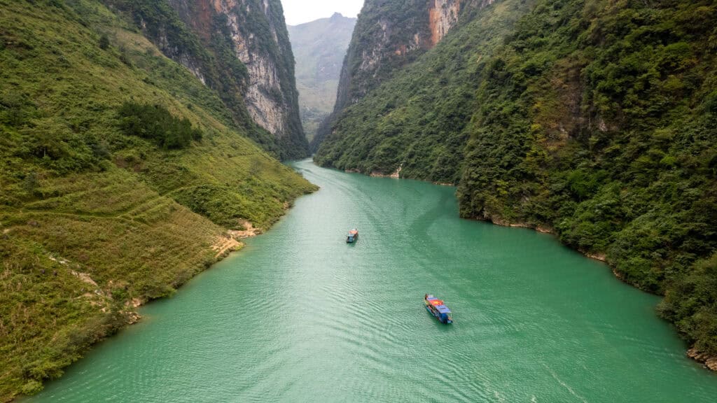 A turquoise river winds between steep, green mountains. Three boats travel along the water, creating gentle ripples. The landscape appears lush and serene under a cloudy sky.