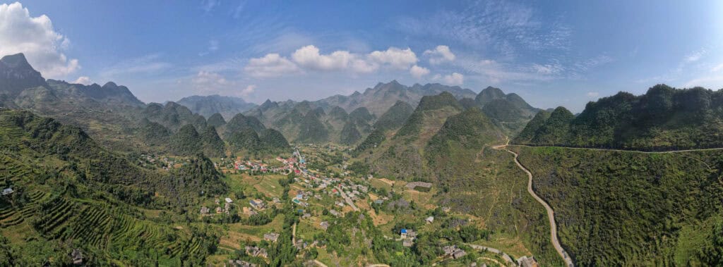Aerial view of a valley town surrounded by green mountains, terraced fields, winding roads, and clear blue sky with scattered clouds.