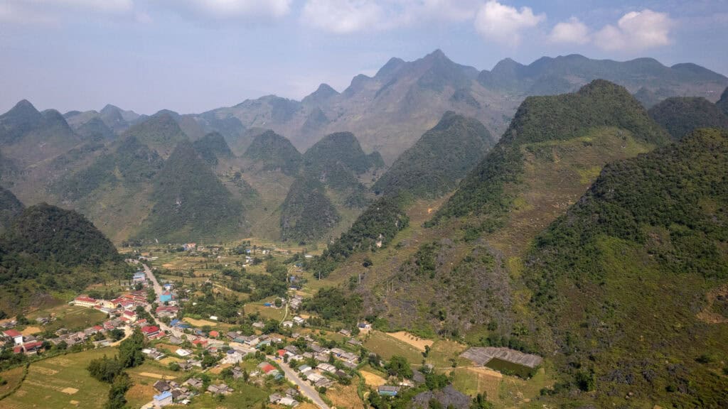 Aerial view of a small village nestled among green, cone-shaped mountains under a partly cloudy sky, with fields and houses scattered throughout the valley.