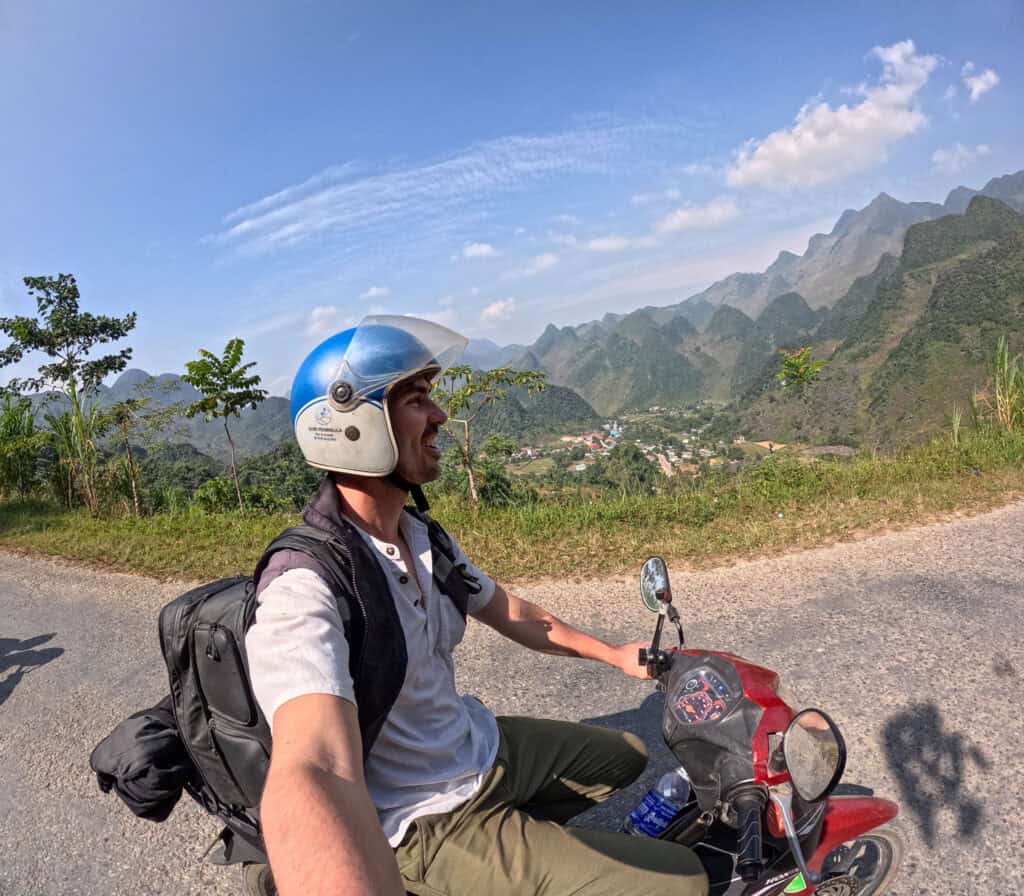A person wearing a helmet and backpack rides a red motorbike on a road with green mountains, blue sky, and scattered clouds in the background. The person is smiling and holding a camera for a selfie.
