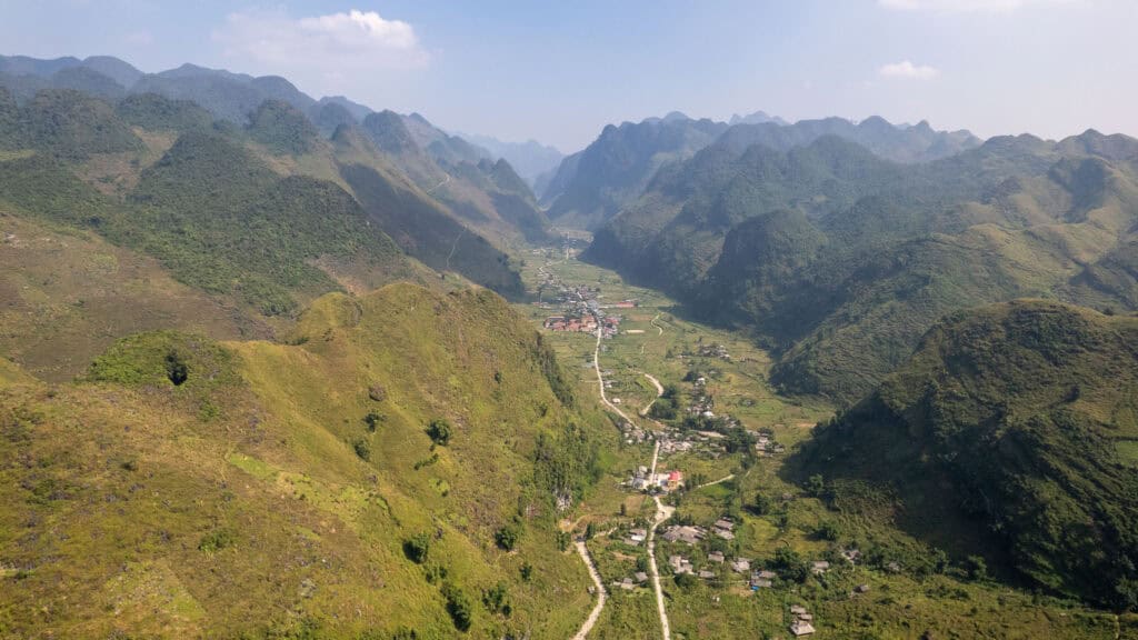 A narrow valley with a winding road runs between tall, green mountains under a blue sky with scattered clouds. Small houses and fields are visible along the valley floor.