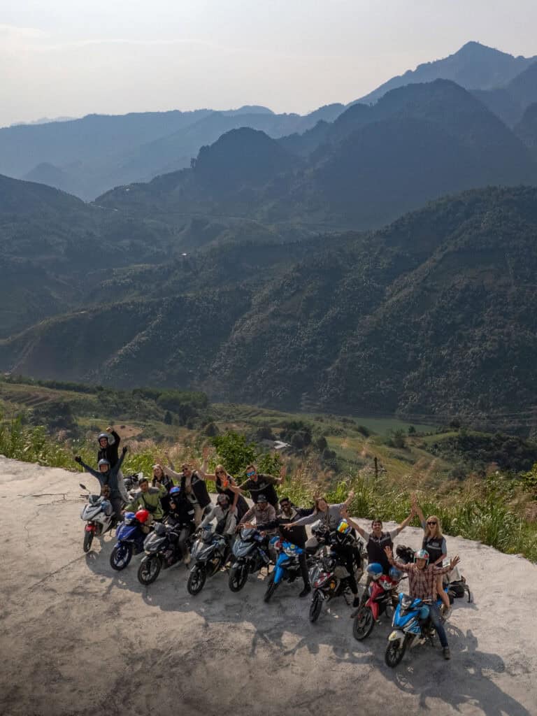 A group of people on motorbikes pose together on a mountain road, with lush green hills and layered mountains in the background under a hazy sky.