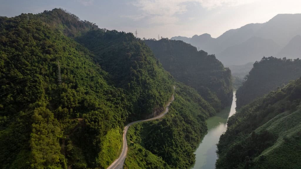 A winding road cuts through lush green hills next to a calm river, with mountains fading into the distance under a partly cloudy sky.
