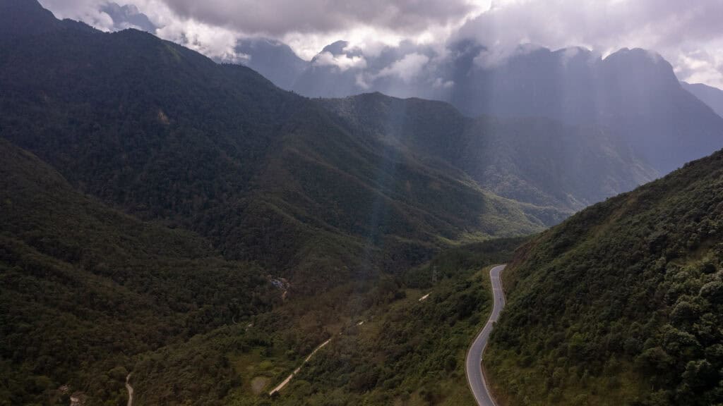 A winding road cuts through the lush green mountains of Tram Ton Pass under a cloudy sky, with rays of sunlight streaming down and creating a dramatic, scenic landscape.