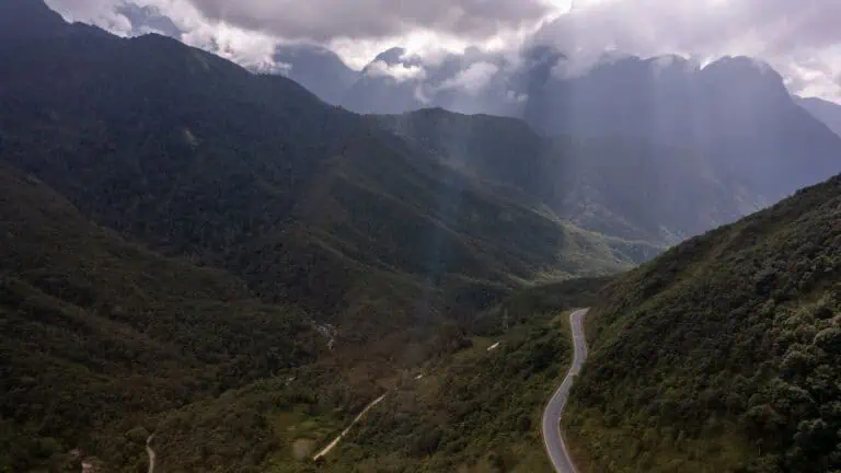 A winding road cuts through the lush green mountains of Tram Ton Pass under a cloudy sky, with rays of sunlight streaming down and creating a dramatic, scenic landscape.