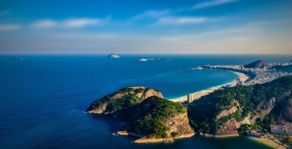 Aerial view of a coastal city with green hills, sandy beach curves, blue ocean, distant islands, and partly cloudy sky—capturing the stunning landscape that surrounds the famous Pedra da Gávea hike, widely considered the best hike in Rio de Janeiro.