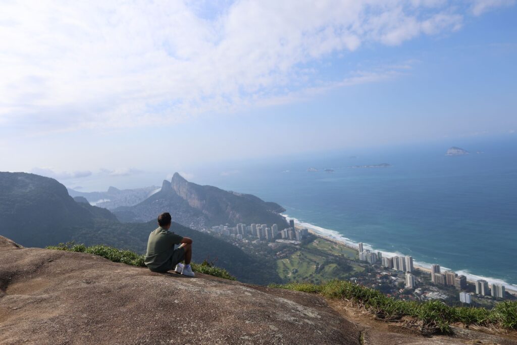 A person sits on a rocky ledge overlooking a coastal city with mountains, tall buildings, and a blue ocean beneath a partly cloudy sky, capturing the breathtaking view after completing the best trail in Rio de Janeiro—the rewarding hike to Morro Dois Irmãos.