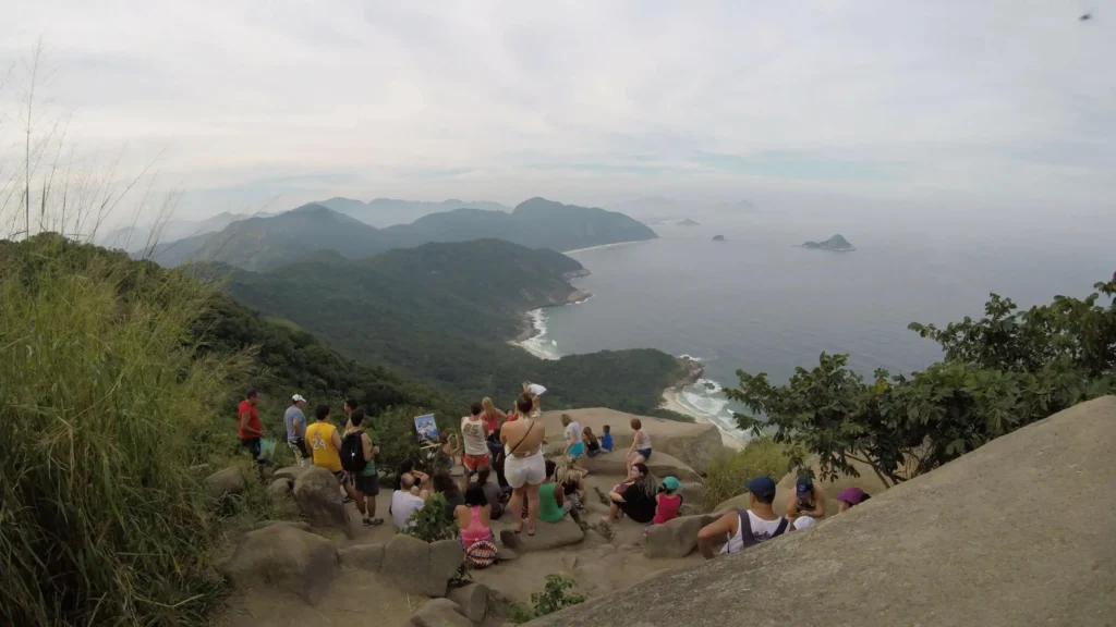 A group of people gather on a rocky viewpoint overlooking lush green mountains and a coastline with waves and distant islands under a cloudy sky, capturing the breathtaking beauty of the famous Morro Dois Irmãos after an unforgettable hike in Rio de Janeiro.